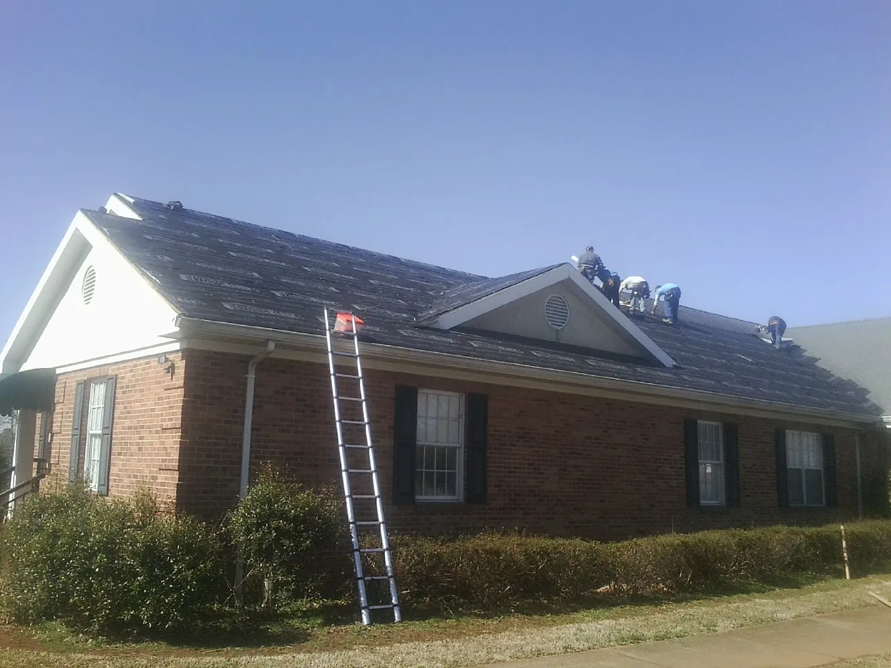 Crew tearing shingles off a brick ranch roof with a ladder set — Metal Roof Repair work by our crew in Little Chute