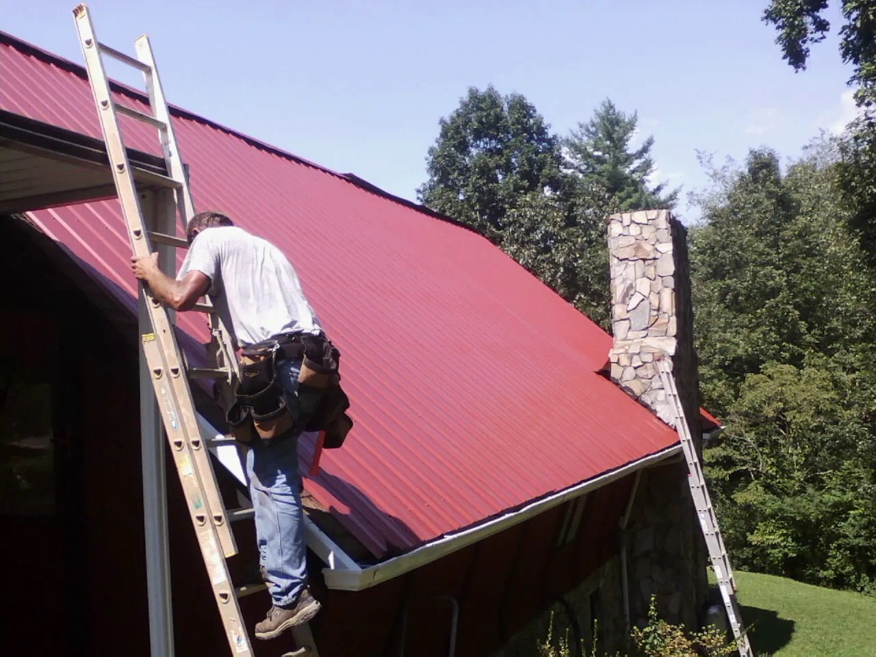 Roofer on a ladder beside a red metal roof and stone chimney — Soffit & Fascia Repair by Roofing Brothers in Little Chute